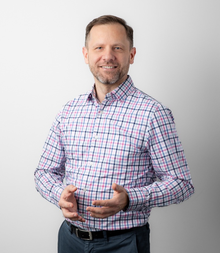 Portrait of a smiling middle-aged man in a pink-and-blue checkered shirt, standing against a neutral background with hands gesturing.