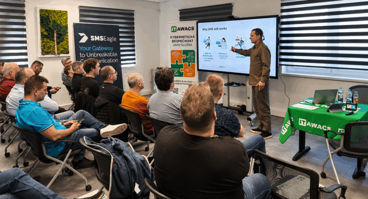 Presenter in brown shirt explains slide to a seated audience in a training room; screen shows 'Why SMS still works' and IT AWACS banners are visible.