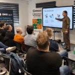 Presenter in brown shirt explains slide to a seated audience in a training room; screen shows 'Why SMS still works' and IT AWACS banners are visible.