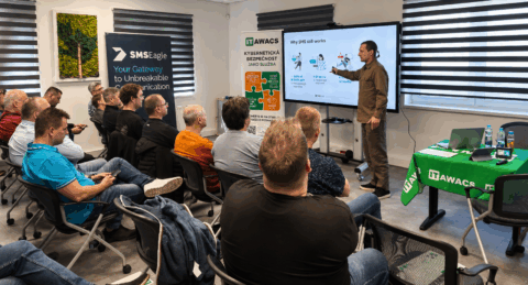 Presenter in brown shirt explains slide to a seated audience in a training room; screen shows 'Why SMS still works' and IT AWACS banners are visible.