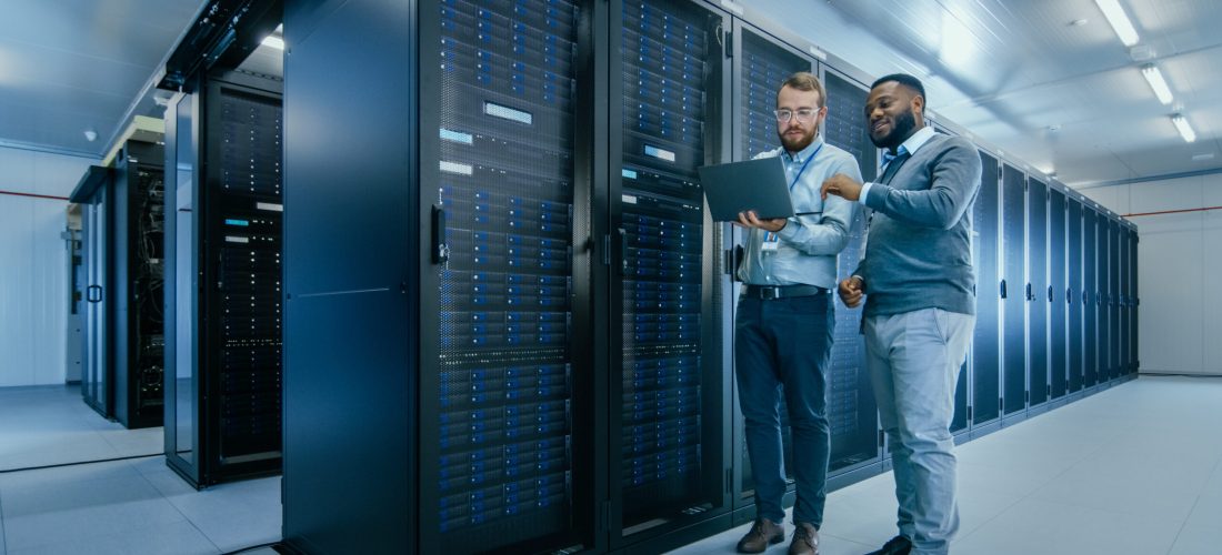 Bearded IT Technician in Glasses with a Laptop Computer and Black Male Engineer Colleague are Talking in Data Center while Working Next to Server Racks. Running Diagnostics or Doing Maintenance Work.